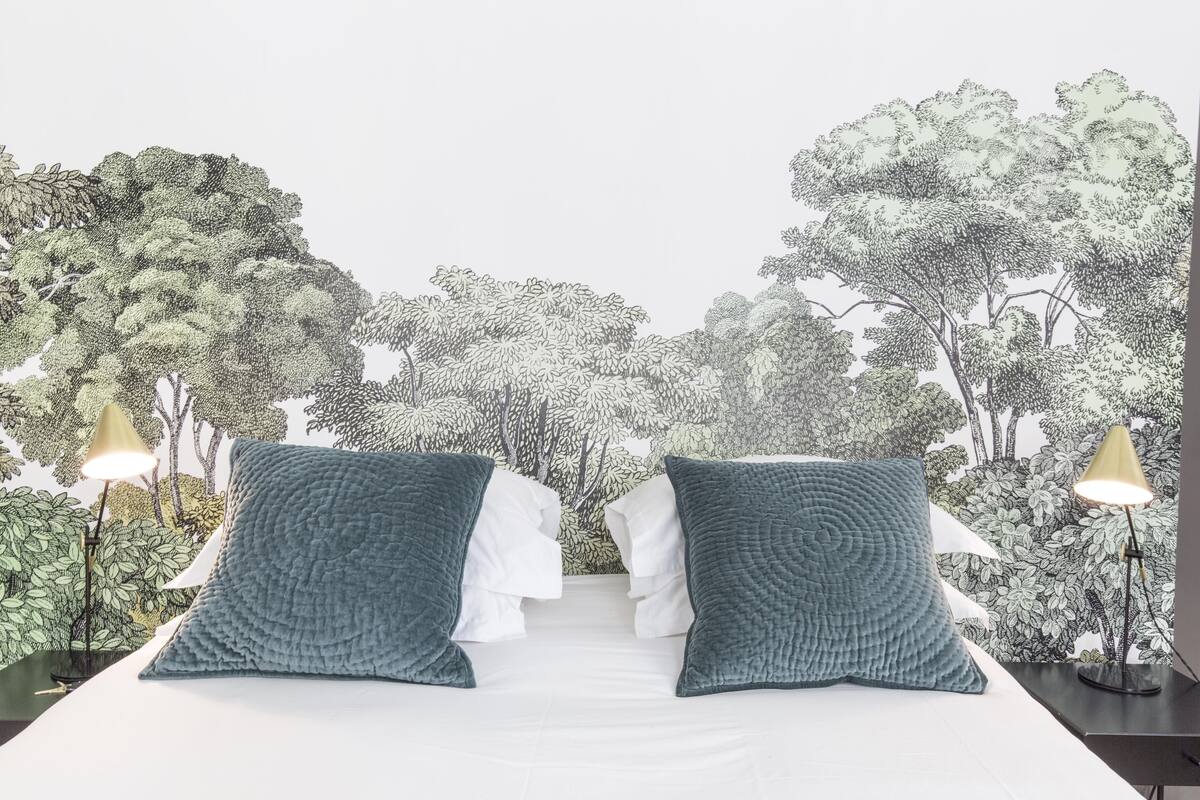 Ein Bett mit zwei Kissen, ein Nachttisch mit Lampe und eine Wand mit einem Wandbild zum Thema Natur.