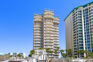 A multi-story hotel with balconies, located near a beach with palm trees.