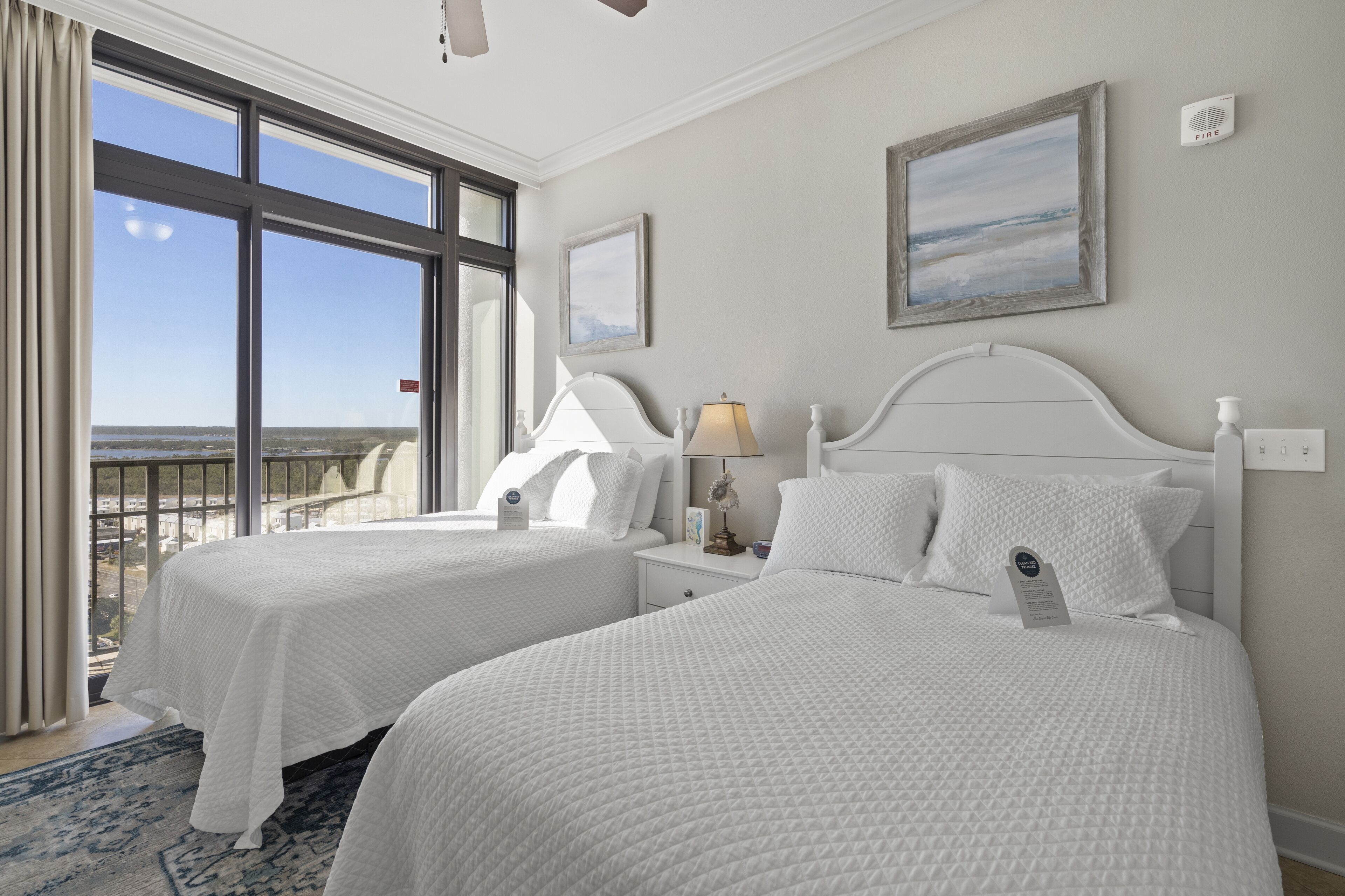 A hotel room with two beds, a large window with a view, a ceiling fan, and a fire alarm.