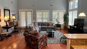 A spacious living room with a large sectional sofa, a floral armchair, and a television. There are multiple windows with blinds, a large plant, and a patterned rug.