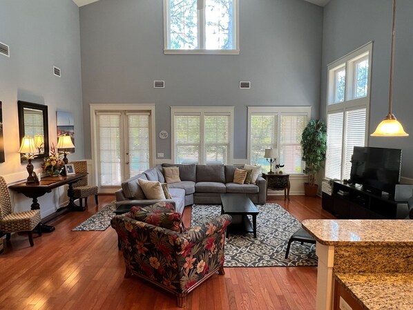 A spacious living room with a large sectional sofa, a floral armchair, and a television. There are multiple windows with blinds, a large plant, and a patterned rug.