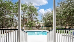 Zona de piscina con cerca blanca, cubierta de madera y árboles al fondo.