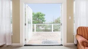Habitación con puerta de cristal corredera que da a una terraza exterior con barandal blanco y vistas a árboles y edificios.