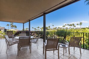A covered outdoor seating area with wicker chairs and a round table, overlooking a tropical landscape with palm trees and a clear blue sky.