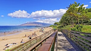 A beach with a wooden boardwalk, people sunbathing, and a mountain in the background.