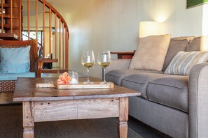 A wooden coffee table with two wine glasses and a flower centerpiece in a living room with a gray sofa and a wooden staircase in the background.