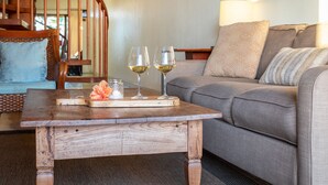 A wooden coffee table with two wine glasses and a flower centerpiece in a living room with a gray sofa and a wooden staircase in the background.