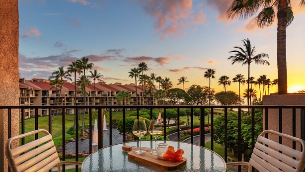 A round glass table with two chairs on a balcony overlooking a resort area with palm trees and a view of the sunset.