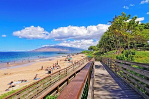 A beach with a wooden boardwalk, people sunbathing, and a mountain in the background.