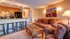 A cozy living room with a sofa, a coffee table, and a kitchen area with wooden cabinets and a refrigerator. - Peak 8 Village Hideaway Condo: On Shuttle Route (Breckenridge)