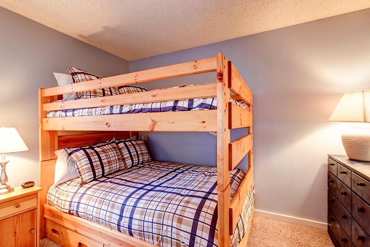 Un lit superposé avec une couvre-lit à carreaux, une table de chevet en bois avec une lampe et une commode avec des tiroirs.