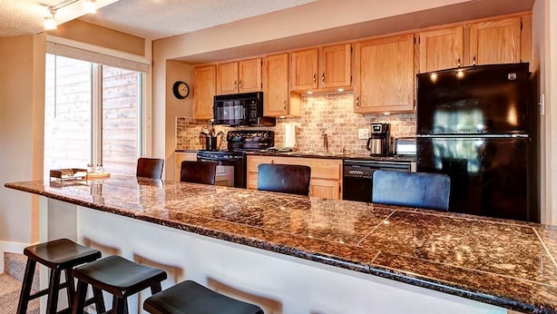 A kitchen with wooden cabinets, a black refrigerator, a microwave, and a granite countertop. - Peak 8 Village Hideaway Condo: On Shuttle Route (Breckenridge)