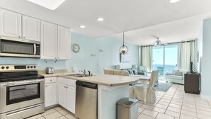 A modern kitchen with white cabinets, a built-in microwave, and a dishwasher. There is a dining area with a table and chairs, and a view of the outdoors through large windows. - Lighthouse 1112 by Liquid Life (Gulf Shores)