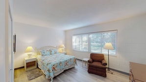 A bedroom with a bed, two bedside tables with lamps, a chair, a window with blinds, and a door. - Atlantic Breeze #2 2 bedrooms, 1 bath (Jekyll Island)