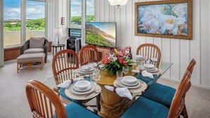 Een eetruimte met een tafel voor zes personen, omringd door rieten stoelen, een groot schilderij en een televisie.