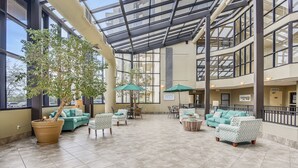 A spacious indoor area with a large potted tree, green and white patterned sofas, and a glass ceiling. - Phoenix 7 1011 | Gulf Views | Indoor, Outdoor Pools | My Beach Getaways (Orange Beach)