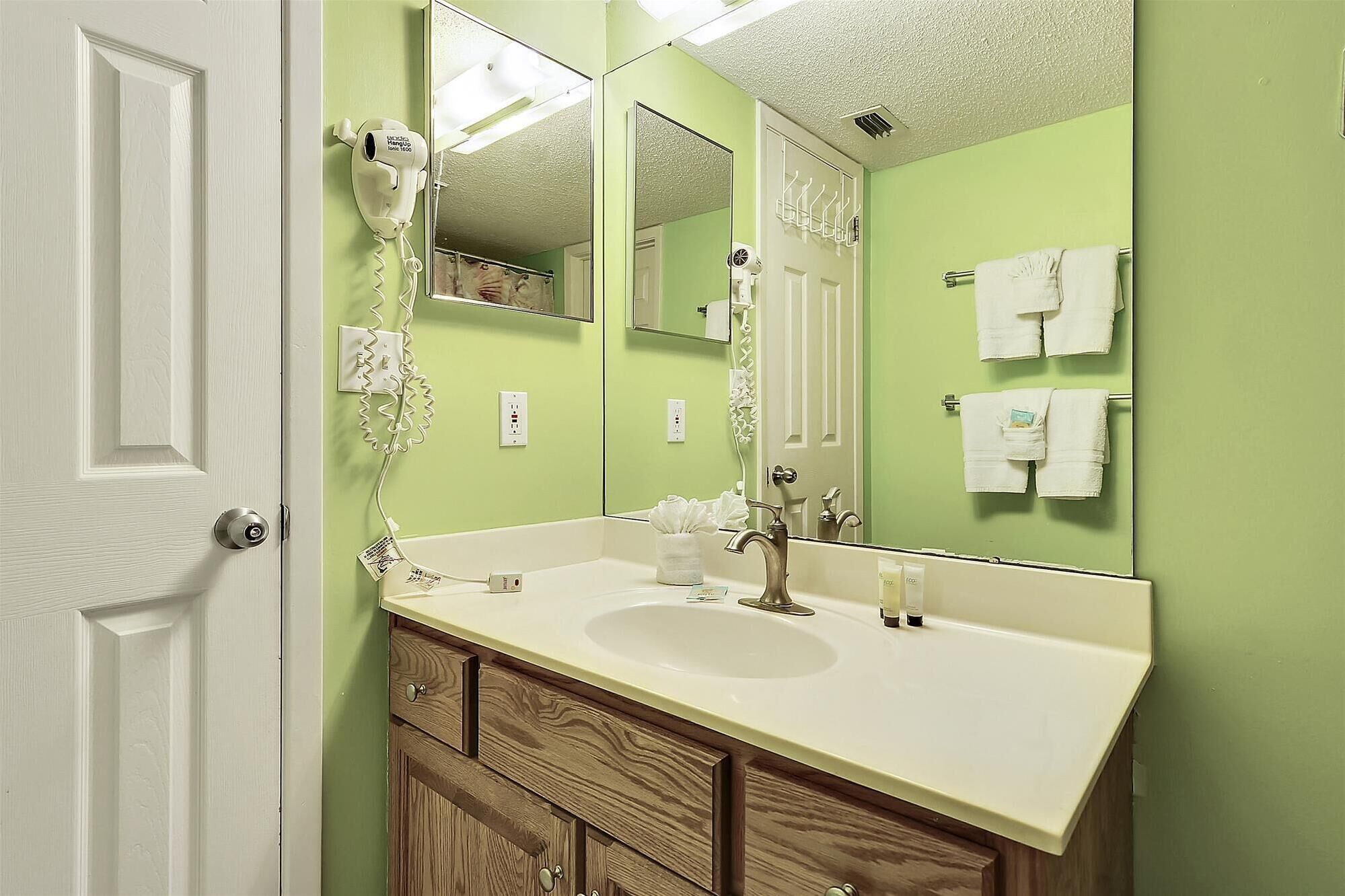 Une salle de bain avec un lavabo blanc, des meubles en bois, un grand miroir et un sèche-cheveux fixé au mur.