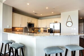 Una cocina moderna con armarios blancos, electrodomésticos de acero inoxidable, una encimera de mármol y una zona con taburetes de bar.
