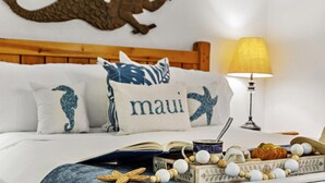A neatly made bed with a wooden headboard, decorative pillows with seahorse and 'maui' prints, a bedside lamp, and a tray with snacks and a book. - Kahana Villas E308  Charming Condo (Lahaina)