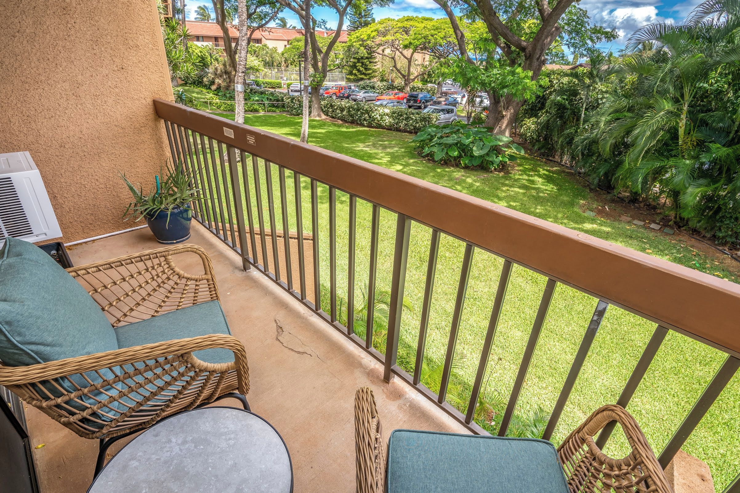 A balcony with wicker furniture, a potted plant, and a view of a well-maintained lawn and parked cars.
