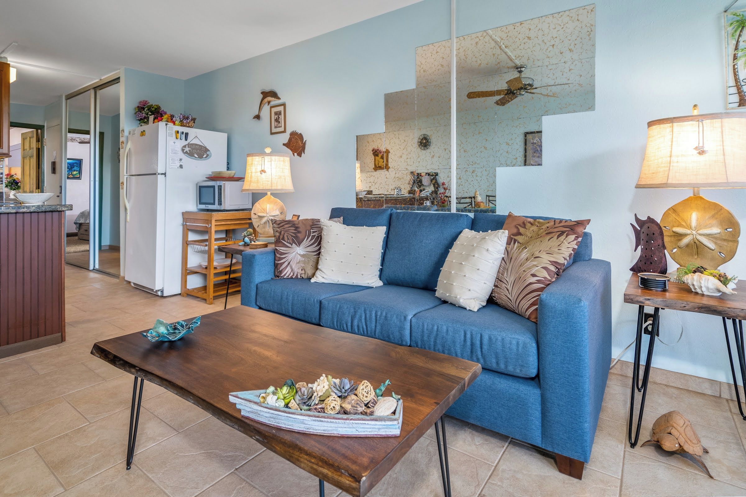 A living room with a blue sofa, wooden coffee table, and a kitchen area with a refrigerator and microwave.