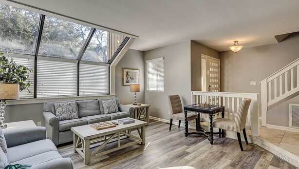 A cozy living room with a sofa, coffee table, and a chess table. There are windows with blinds, a staircase, and a door with a window. - 119 Beachwalk - 5 Minute Walk to the Beach - Enjoy a Pet Friendly getaway! (Hilton Head Island)