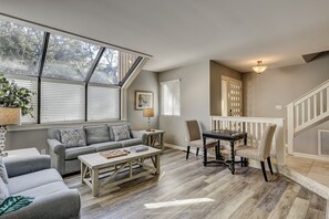 A cozy living room with a sofa, coffee table, and a chess table. There are windows with blinds, a staircase, and a door with a window. - 119 Beachwalk - 5 Minute Walk to the Beach - Enjoy a Pet Friendly getaway! (Hilton Head Island)