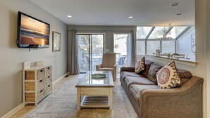 A living room with a sofa, a coffee table, a TV mounted on the wall, and a window with a view of the outside. - 119 Beachwalk - 5 Minute Walk to the Beach - Enjoy a Pet Friendly getaway! (Hilton Head Island)