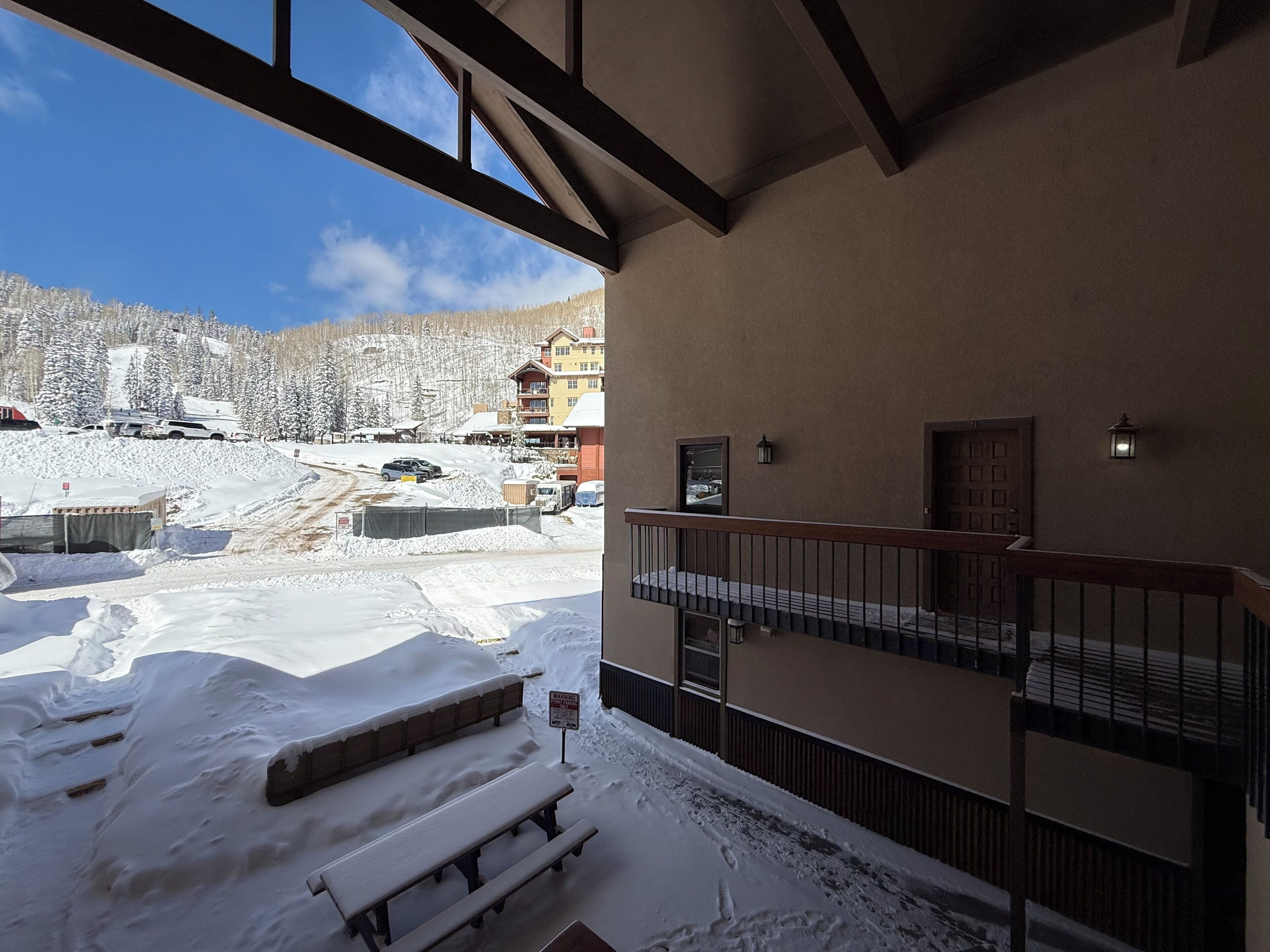 A snow-covered outdoor area with a wooden bench and a warning sign, a balcony with a railing, and a view of a ski resort in the background.