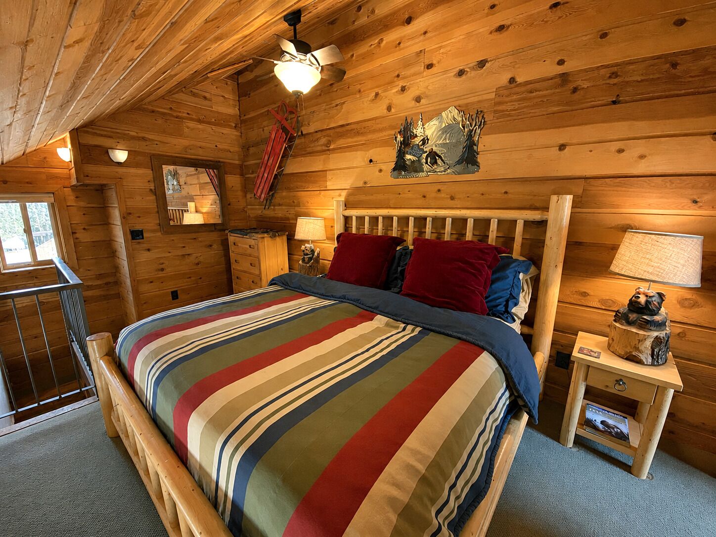 Une chambre dans un chalet en bois, comprenant un lit, une table de chevet, une lampe, une fenêtre et un ventilateur de plafond.