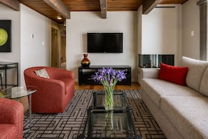 A living room with a sofa, a red armchair, a glass coffee table, a black TV, and a vase with flowers.
