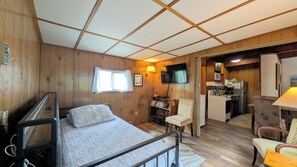 Une pièce de taille modeste comprenant un lit, une chaise, un petit bureau, un téléviseur et un coin cuisine.