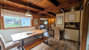 L’intérieur d’un chalet en bois comprend une table à manger, des bancs, un coin cuisine et une chambre à coucher, visible à travers une porte ouverte.