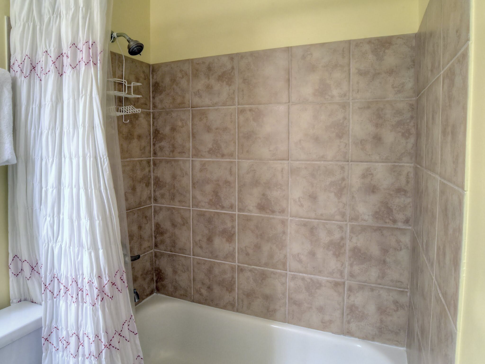 A bathroom with a bathtub, a shower area with beige tiles, and a white shower curtain with red stitching.