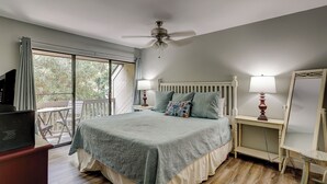 Un dormitorio con cama, ventilador de techo, mesita de noche con lámpara, cómoda y un amplio ventanal.
