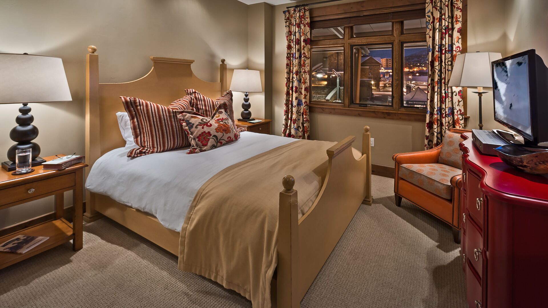 Een hotelkamer met een bed, twee nachtkastjes, een televisie, een rode ladekast en een raam met gordijnen.