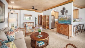 A spacious living room with a sofa, a coffee table, and a TV. There is a kitchen area with wooden cabinets and a refrigerator. - Poipu Sands 532 Panoramic Oceanview  By Suite Paradise (Koloa)