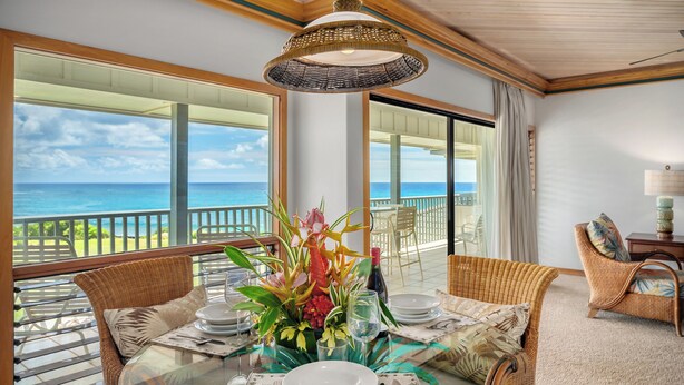 A dining area with a glass table, wicker chairs, and a view of the ocean. - Poipu Sands 532 Panoramic Oceanview  By Suite Paradise (Koloa)