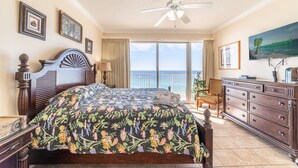 A bedroom with a large bed, a ceiling fan, a dresser, and a view of the ocean.