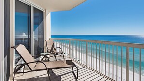 A balcony with two lounge chairs and a table, overlooking a beach with clear blue water.