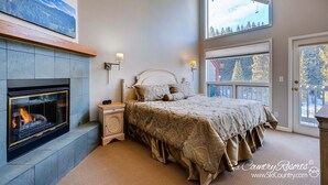 A bedroom with a fireplace, a bed with a patterned bedspread, and a view of trees through the windows.