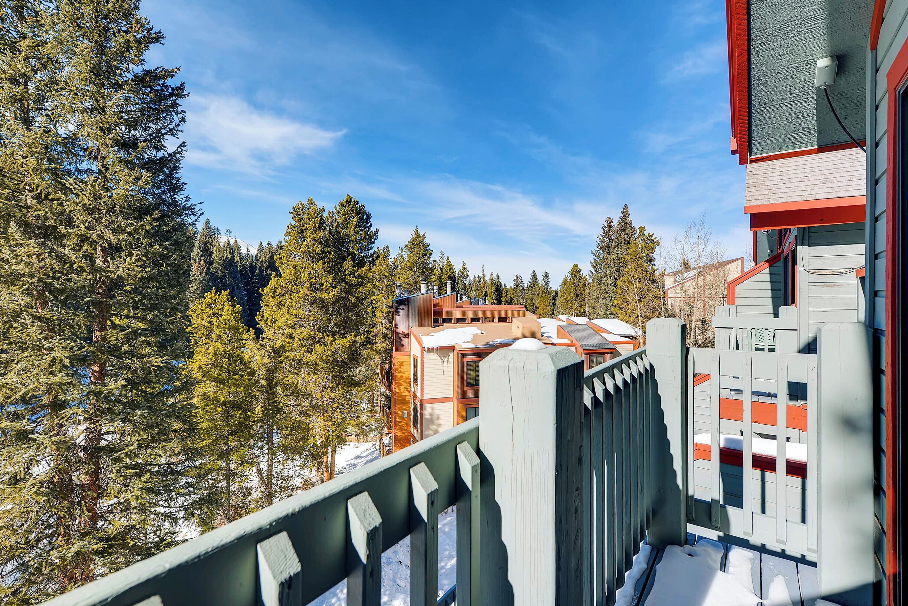 A view from a balcony with snow on the ground, overlooking a building with multiple stories and a forested area.