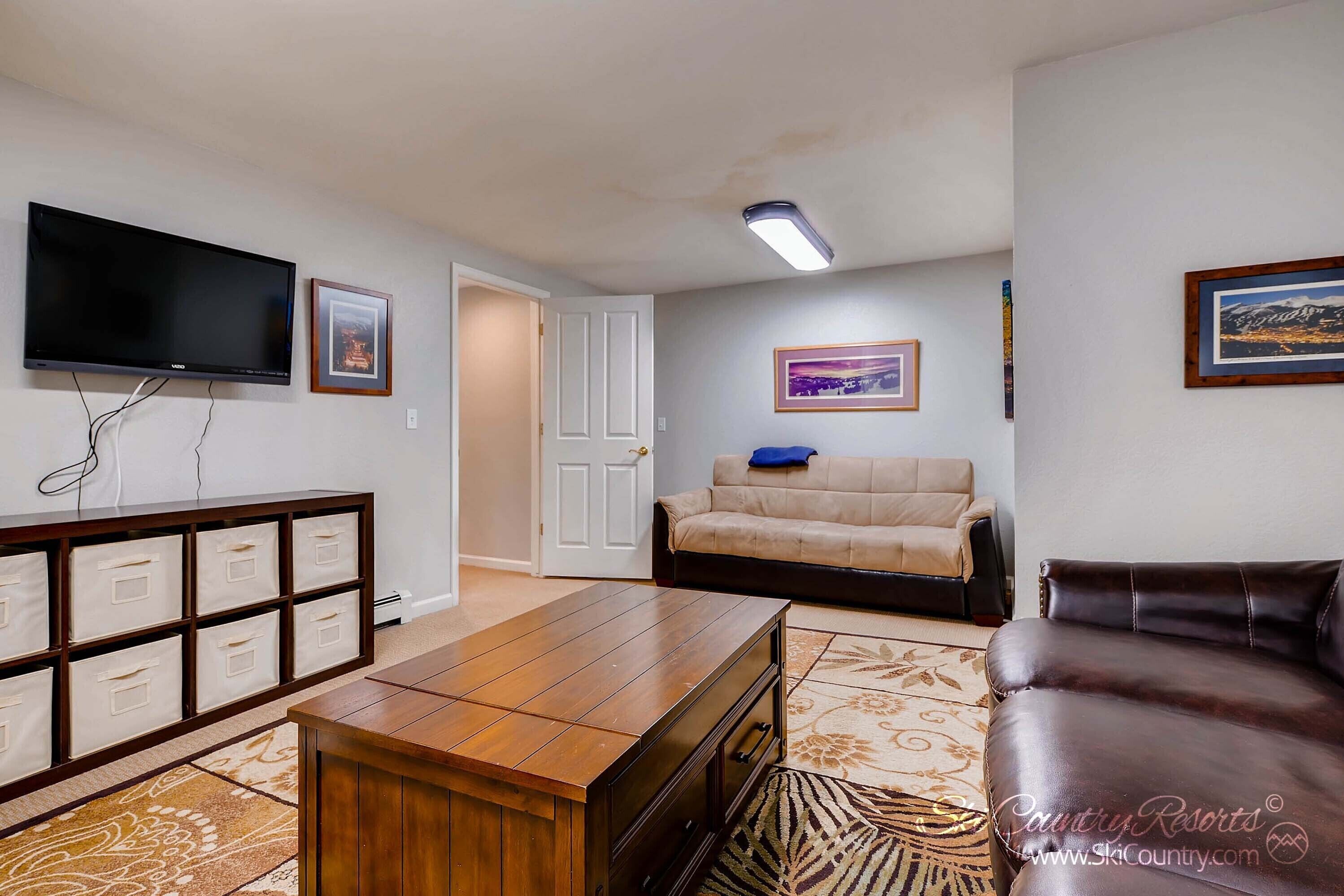 A living room with a flat-screen TV, a wooden coffee table, a beige sofa, a brown leather sofa, a patterned rug, and a storage unit with multiple drawers.