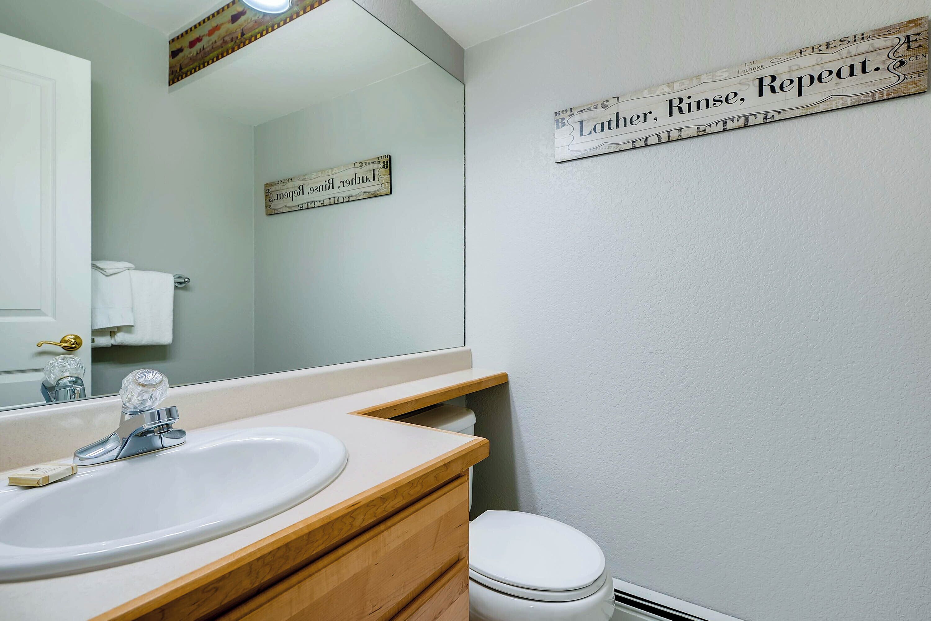 Une salle de bain avec un lavabo blanc, un miroir et des toilettes. Il y a une affiche sur le mur qui indique : « Savonner, rincer, répéter ».