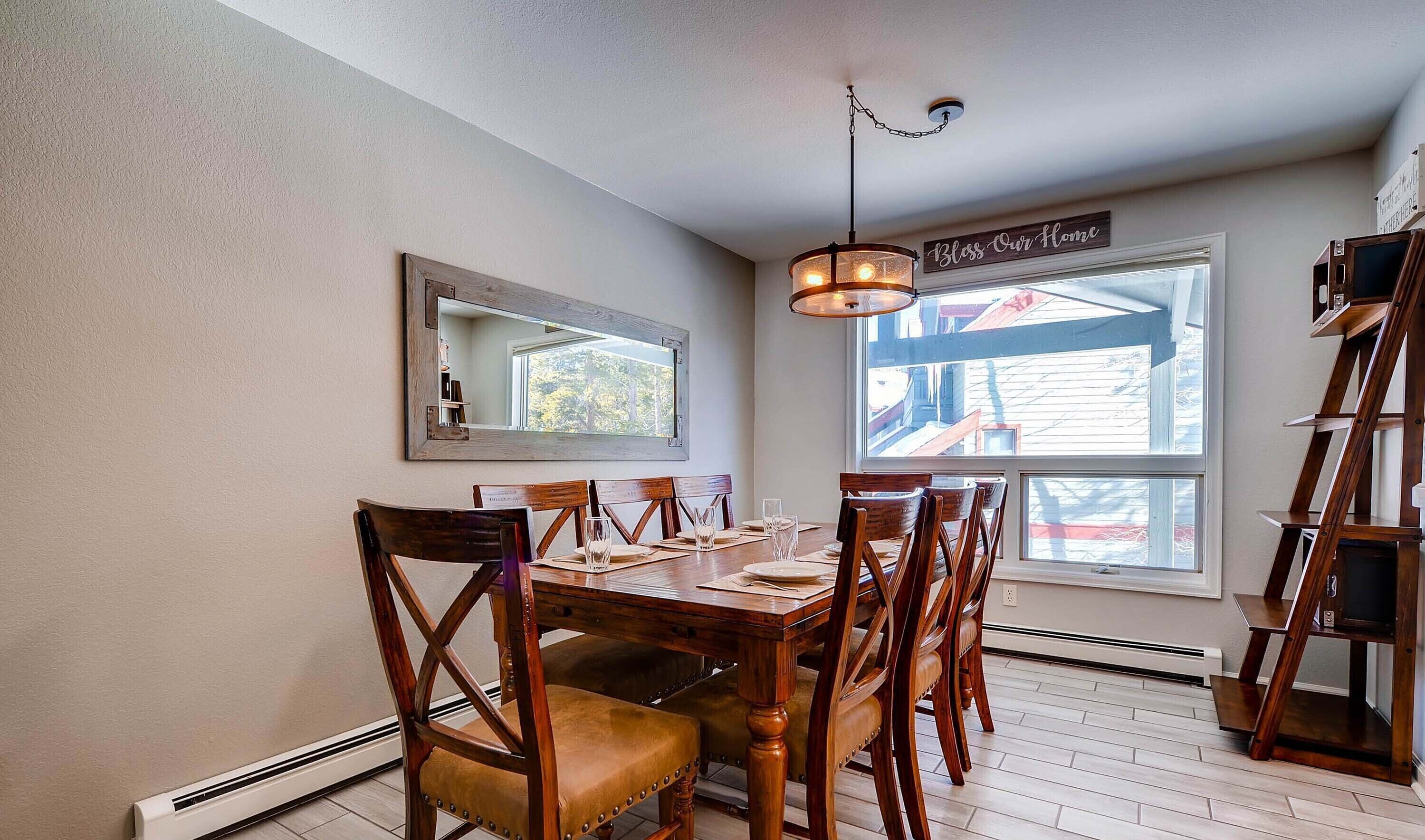 Un'area pranzo con un tavolo e sedie in legno, un lampadario a sospensione, un grande specchio e una finestra vista su un edificio con tetto rosso.