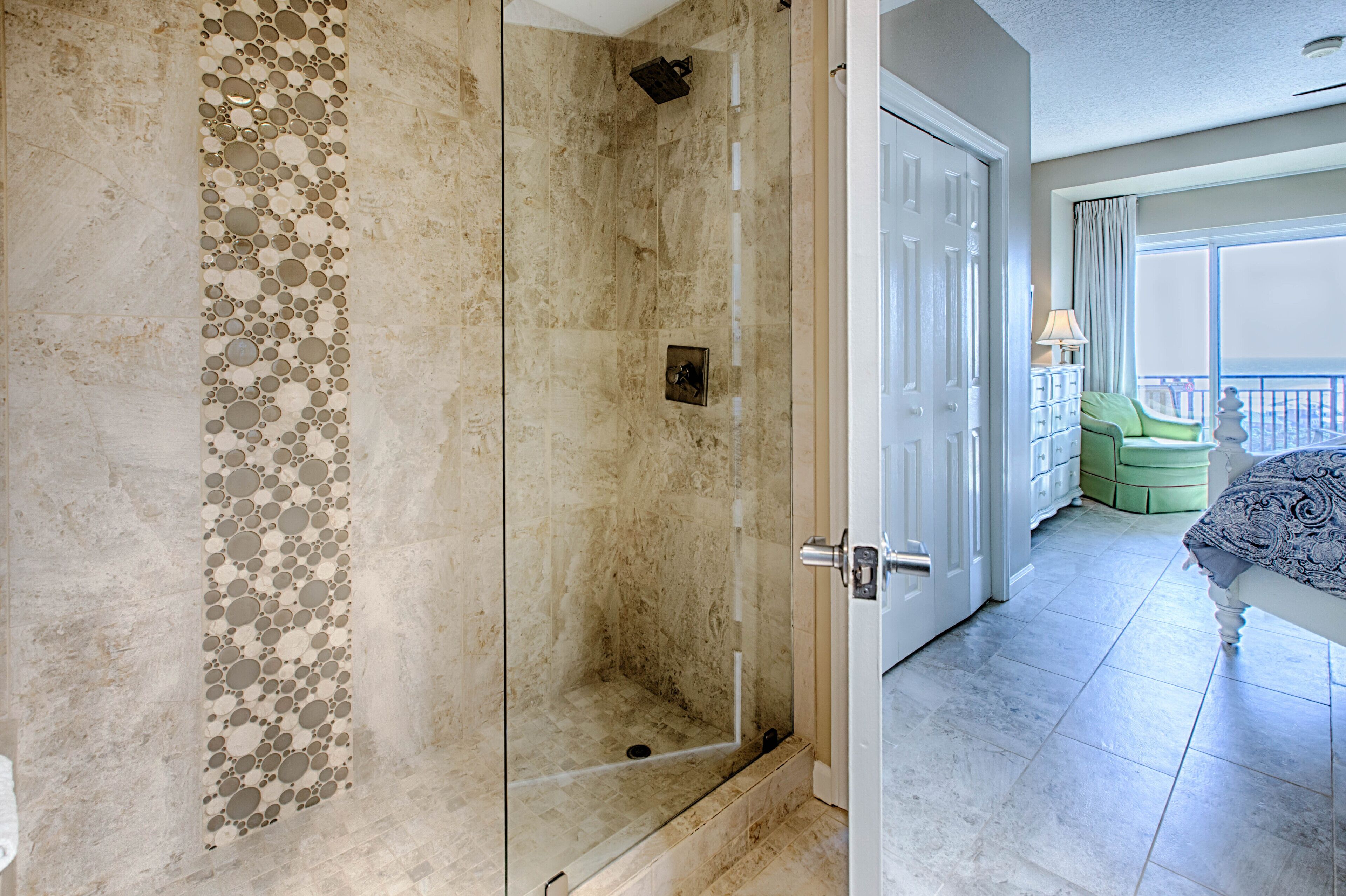 A modern bathroom with a glass-enclosed shower, a marble wall with a circular pattern, and a view of a balcony with a green chair.