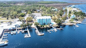 An aerial view of a coastal marina with multiple docks and boats, surrounded by lush greenery and a large building with a blue facade. - Waterfront, RIVERS EDGE CONDO #1B-BLDG 2 PRIVATE DOCK and ASSIGNED BOAT SLIP! (Steinhatchee)