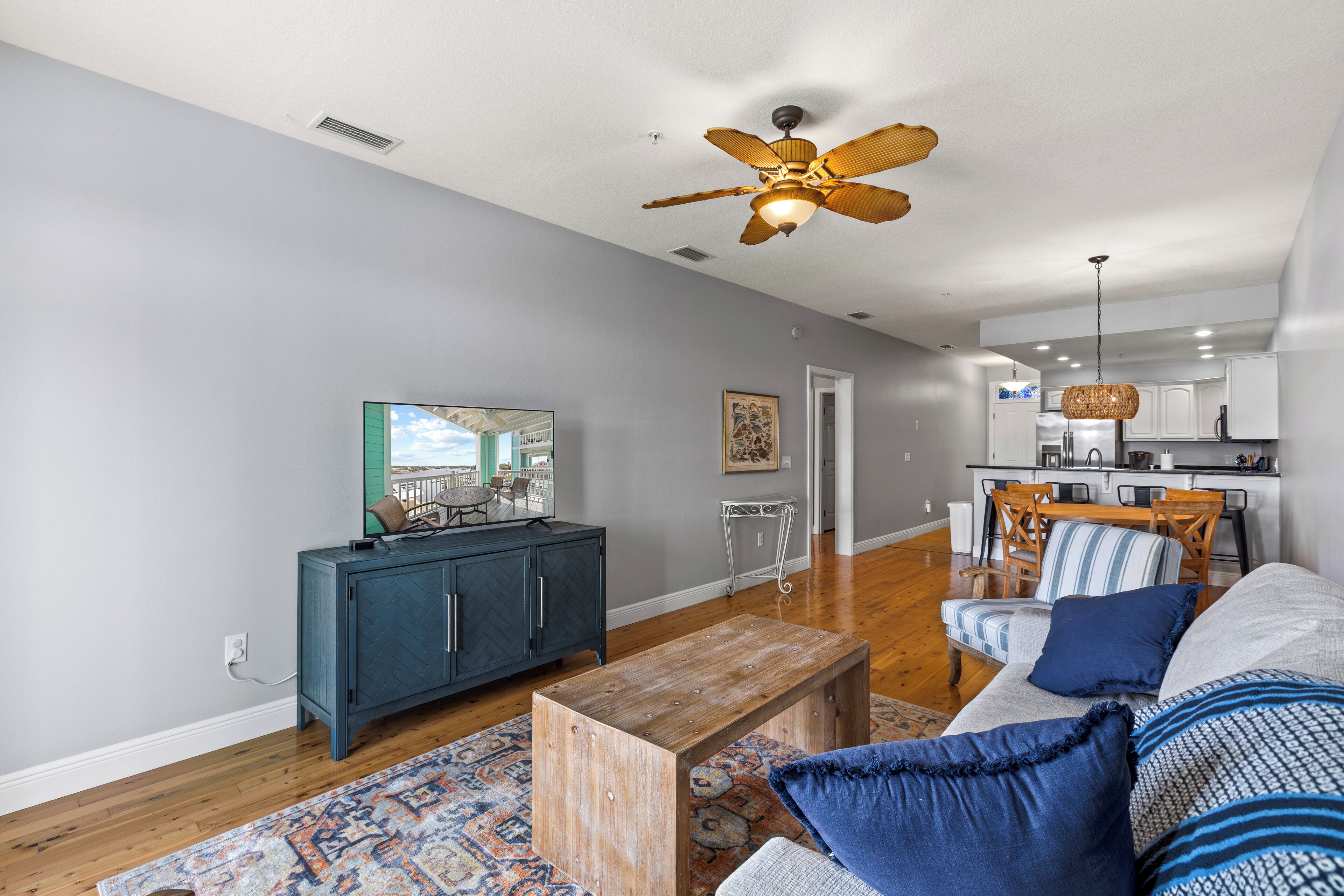 A living room with a sofa, coffee table, and a television. There is a kitchen in the background with a dining area.