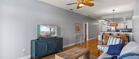A living room with a sofa, coffee table, and a television. There is a kitchen in the background with a dining area.