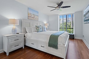 A bedroom with a bed, nightstands, a ceiling fan, a window with a view, and a painting on the wall.